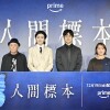青い壁に「人間標本」の大きな文字。その前に3人の男性とその右側に女性1人が立っている。4人の前には、青い蝶の羽をあしらった青いパネルがあり、白い字で「人間標本」と書かれている。