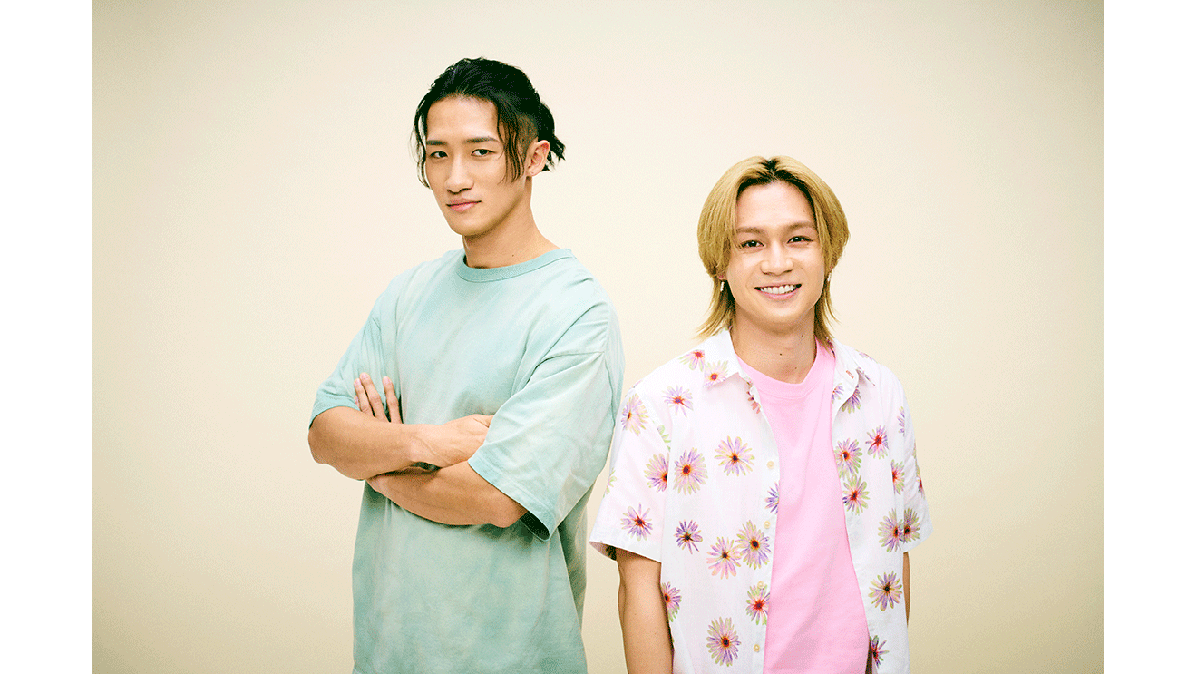 ベージュの背景に並んで立つ2人の男性。左側はTシャツで腕組み、右側はTシャツに花柄のシャツを着用。