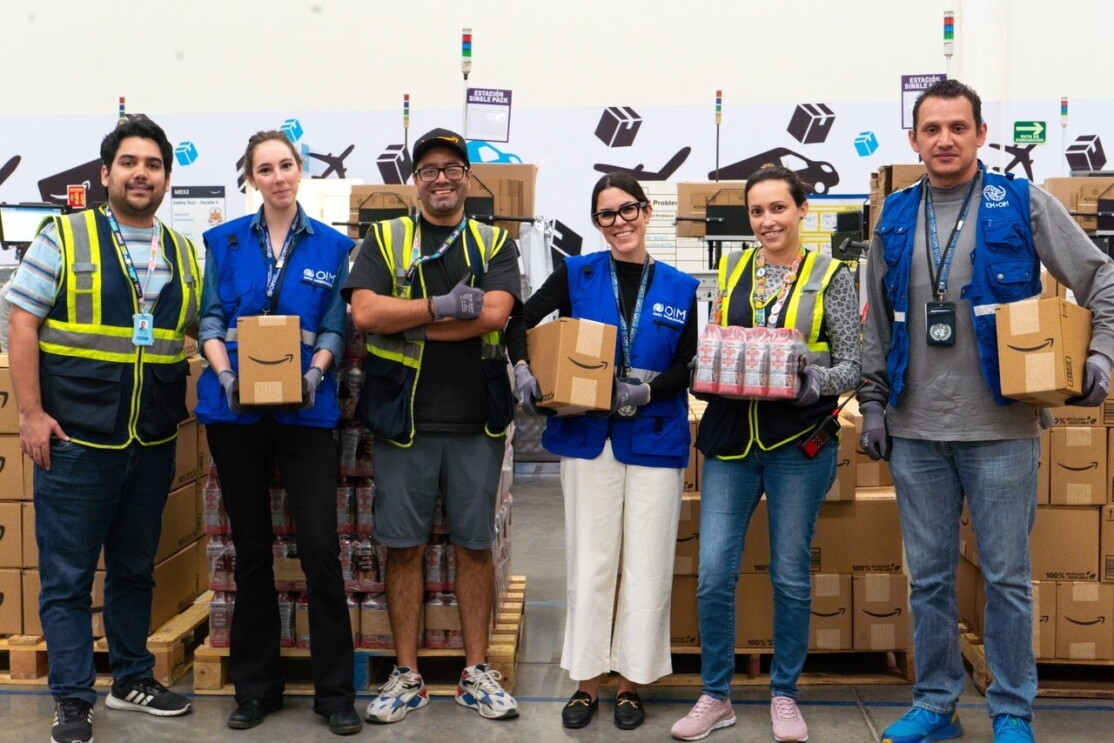 En un centro logístico están tres trabajadores de Amazon posando con tres trab ajamores de la OIM. Detrás hay cajas de Amazon. 