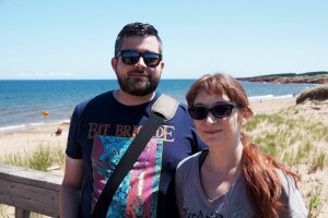 An image of a man and a woman smiling for a photo while standing on a platform in front of a beach.