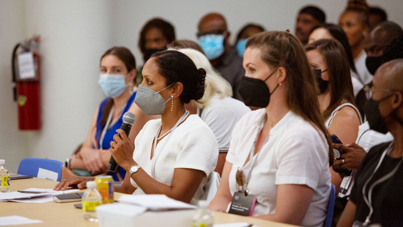 Women sit at a conference table listening to a speaker. They are wearing face masks.