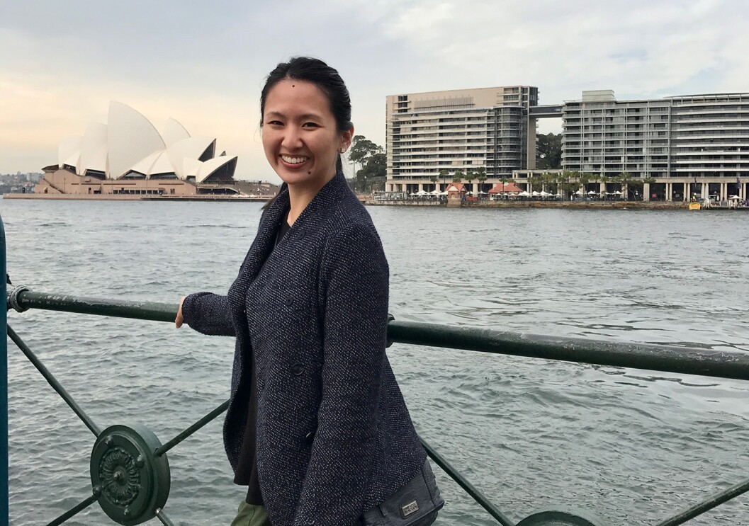 An image of a woman smiling for a phot in front of an ocean bay. The Sydney opera house is in the background behind her.