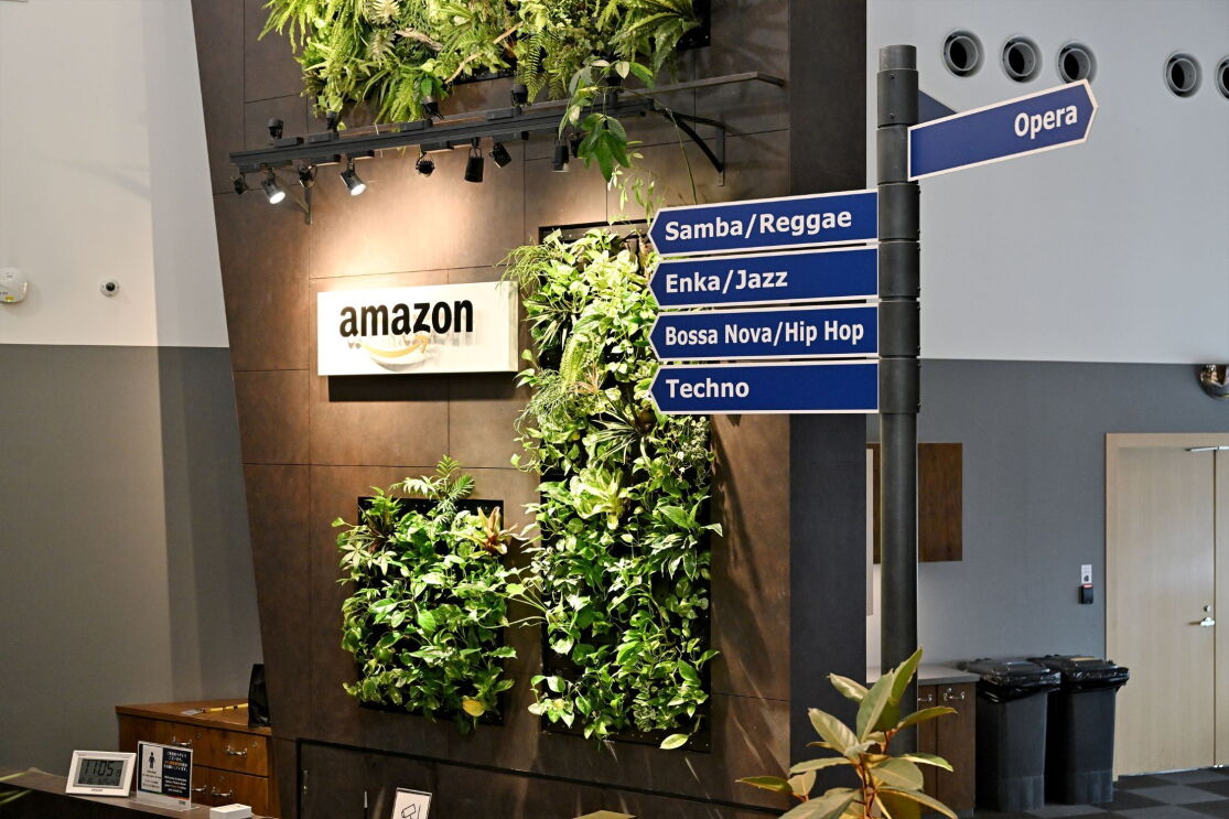 茶色の壁に植物が埋め込まれている。その横にAmazonのロゴ。壁の手前に会議室の方向を示す街路標識のデザインの表示