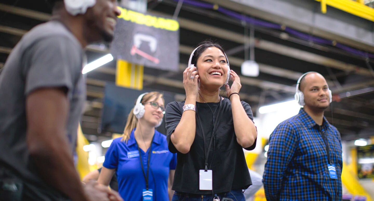 Four people wearing headphones and smiling during an Amazon fulfillment center tour.