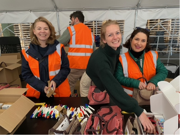 Amazon Mitarbeitende mit orangenen Warnwesten vor einem Tisch mit Buntstiften, Süßigkeiten, Nüssen und Spielsachen, die in die Hilfspakete für Kinder gepackt werden.