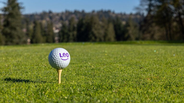 Golf ball with Amazon Leo logo on wooden tee in grass with trees in background