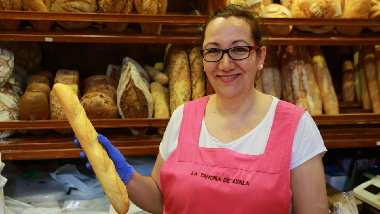 Elena Revuela de La Tahona de Ayala con una barra de pan. Luce delantal rosa con el nombre de su negocio, una camiseta blanca y gafas de pasta negras. Destrás de ella está lleno de panes de diferentes formas y tamaños. 
