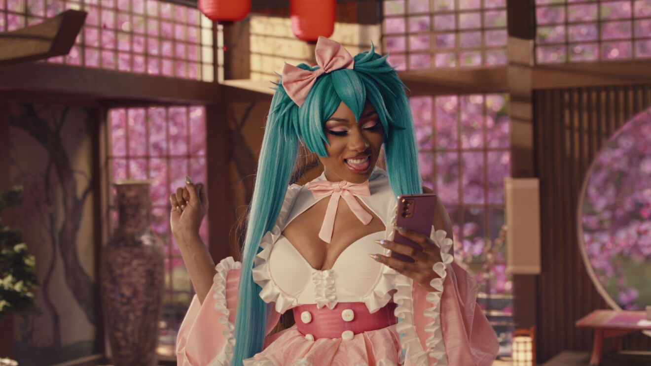 An image of Megan Thee Stallion in a cosplay outfit while smiling and looking at her phone. She is wearing a bright blue wig with long pigtails and an Asian-inspired outfit while standing inside a dojo.