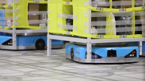グレーの床に、黄色いビニールで覆われ区切られた収納棚。棚の脚は床から浮いていて、下の空間に水色の自動搬送ロボットが格納されている。奥側にも同様の棚とロボット。