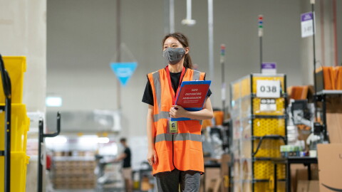 An associate is walking on the floor at Amazon's Fulfillment Center in Singapore with a safety mask on and a file in her hand