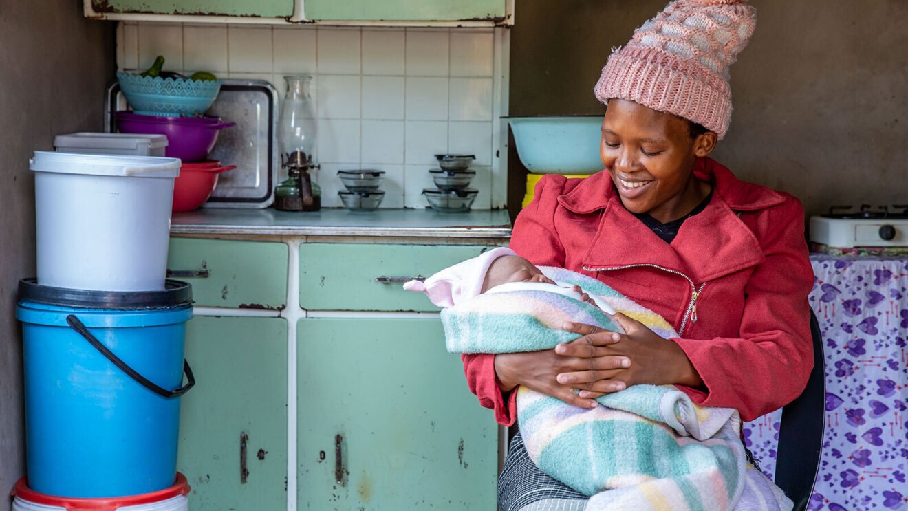 An image of a woman smiling while looking down at a baby she is holding in her arms. The baby is wrapped in a blanket and is wearing a beanie on its head.