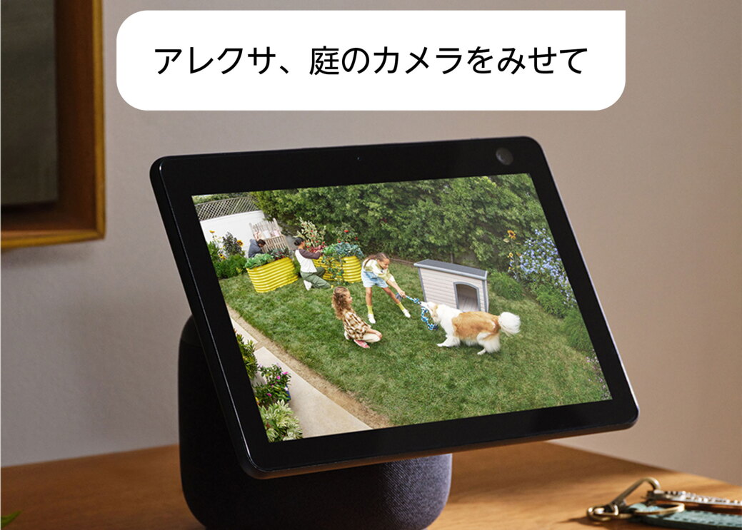 室内の家具の上にEcho Show 10が置かれている。タブレット画面には芝生の庭で犬と遊ぶ2人の子どもたち。製品の上には「アレクサ、庭のカメラをみせて」という文字がある。