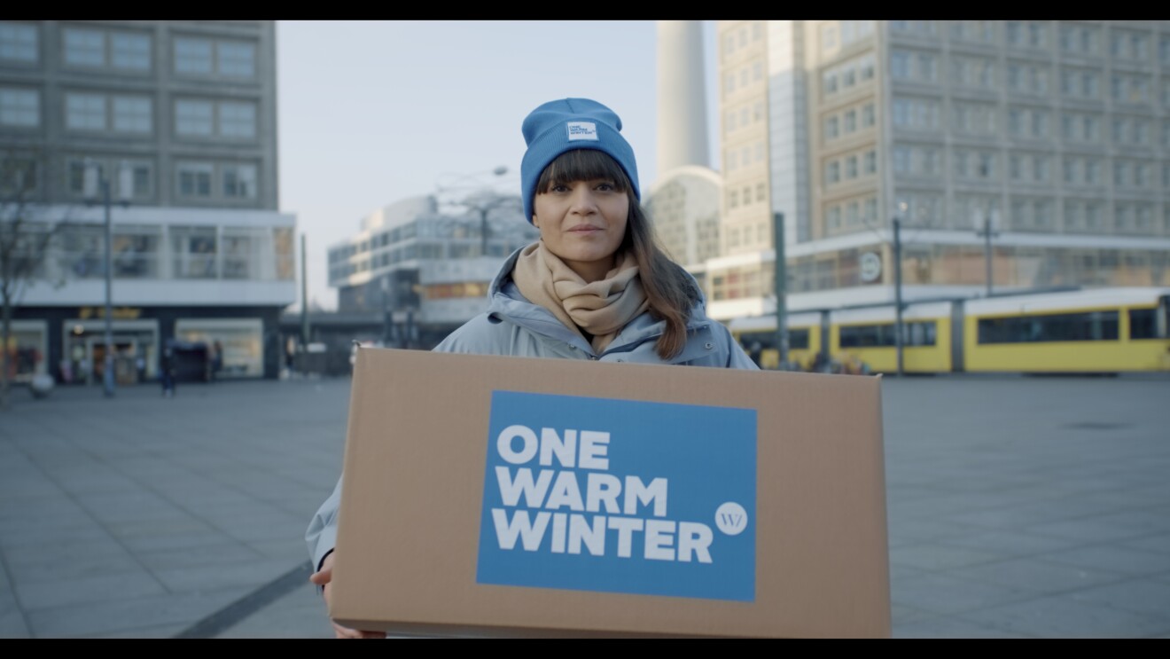 Eine dunkelhaarige Frau mit blauer Mütze schaut in die Kamera. Sie steht auf einem Platz und hat eine Winterjacke an. In der Hand hält sie einen Karton auf dem "One Warm Winter" steht.