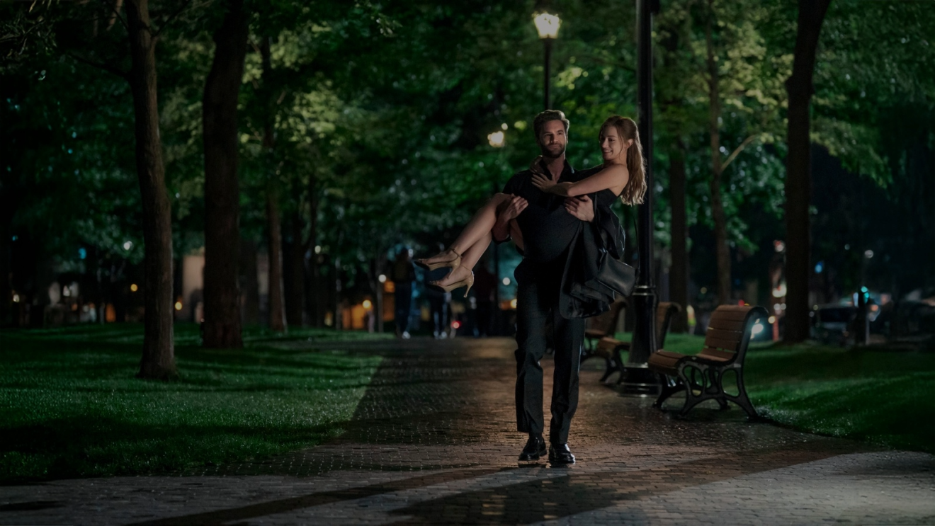 Tom Bateman carrying Lili Reinhart in his arms through the park at night lovingly.
