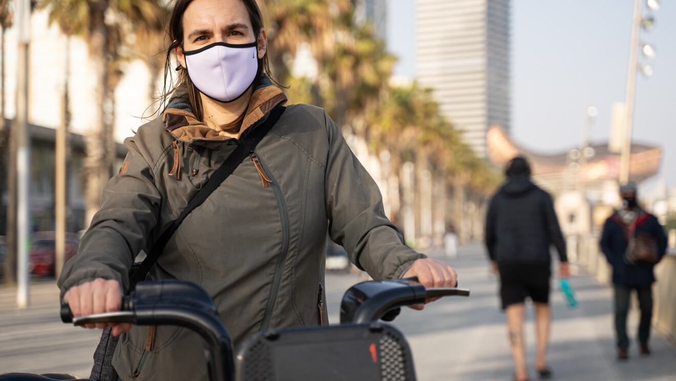Gemma Viladomat, Senior Product Manager para Italia y España  de Amazon. Gemma Viladomat estudió Ingeniería de caminos y en 2016 entró en Amazon.  Gemma en bicicleta en lel paseo marítimo de Barcelona. De fondo las palmeras mientras ella, de pie, agarra el manillar de la bici. Va con una chaqueta puesta. Una cahqueta entre verde y gris y tiene el pelo castaño por el hombro. Lleva una mascarilla blanca.  