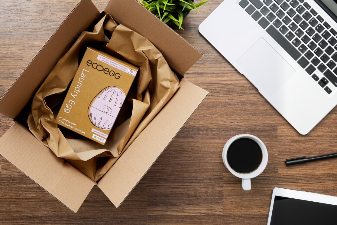 Ein Laundy Egg im Versandkarton von Amazon mit Recycling-Verpackungspapier anstatt mit Plastik Luftpolsterkissen. Daneben liegen ein Laptop, ein Stift, ein Tablet und eine gefüllte Kaffeetasse.