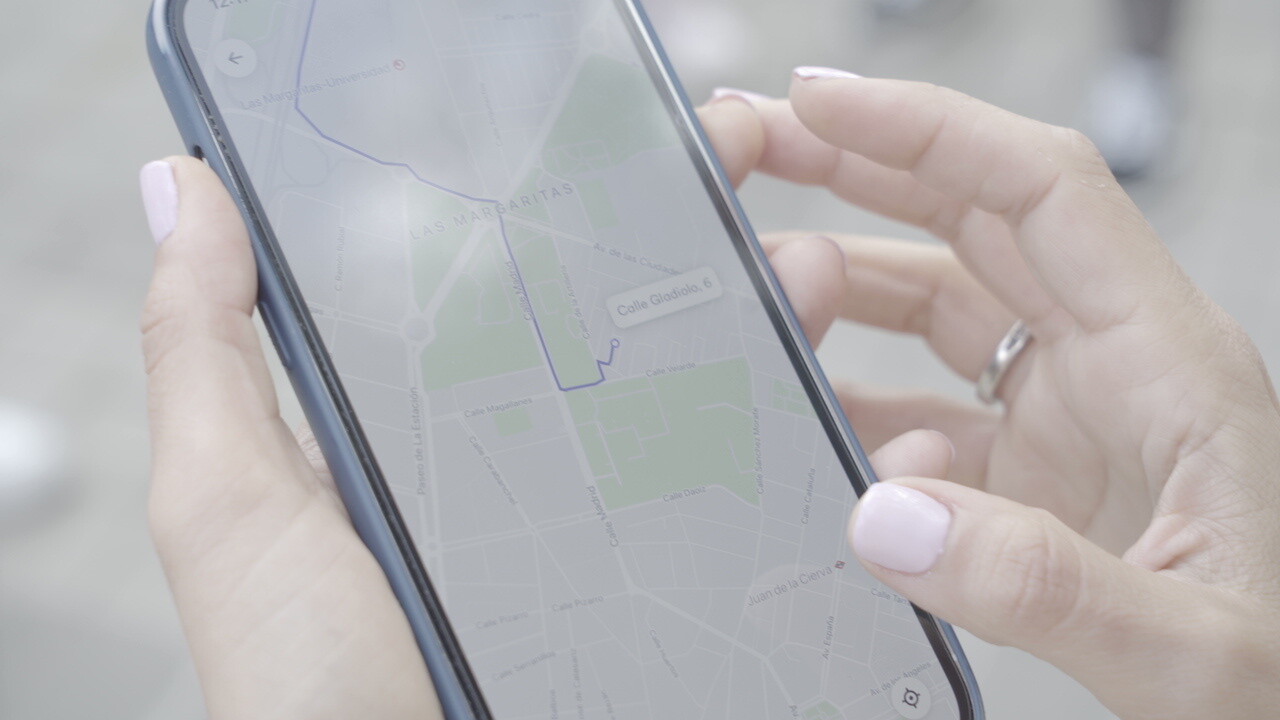 Cabify es la primera empresa europea de movilidad neutra en carbono. Dos manos con la manicura hecha y un anillo de boda en la mano izquierda. Con las dos manos sujeta un teléfono móvil qu een la pantalla tiene abierta la aplicación de Cabify con el recorrorido en color morado hasta llegar al destino deseado. 