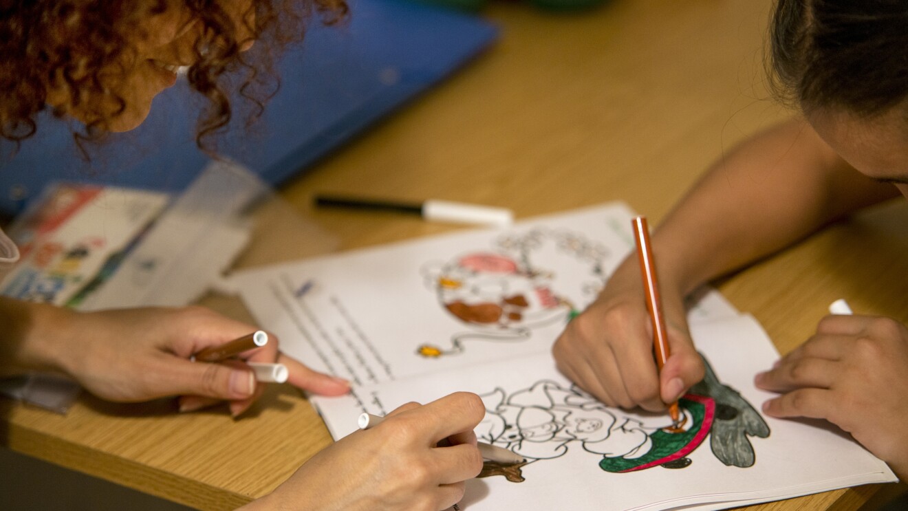 En el primer plano hay una mesa las manos de una niña a la derecha coloreando un cuento con un rotulador marrón. A la izquierda las manos y la cabeza se una madre con el pelo rizado ayudando a pintar a su hija. La mesa es dec color marrón y titne más coloes y una carpeta de color azul.