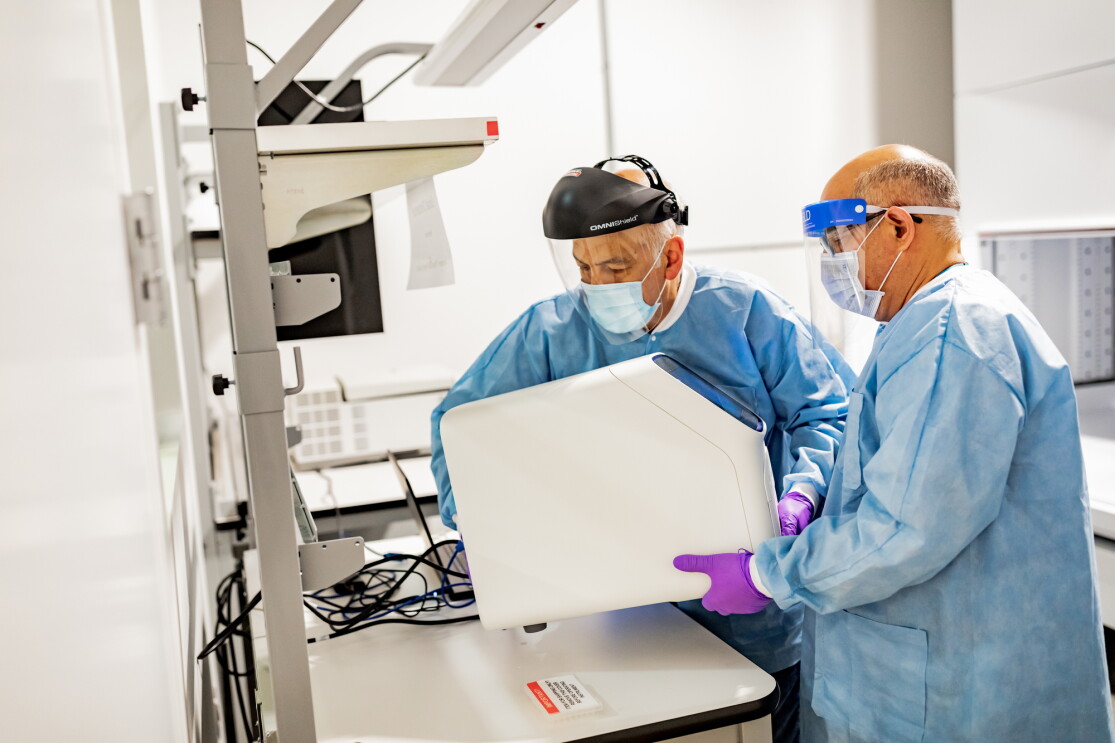Two men in a laboratory setting, wearing protective gear (gloves, mask, face shield, protective clothing) handle a piece of machinery in a laboratory setting. 