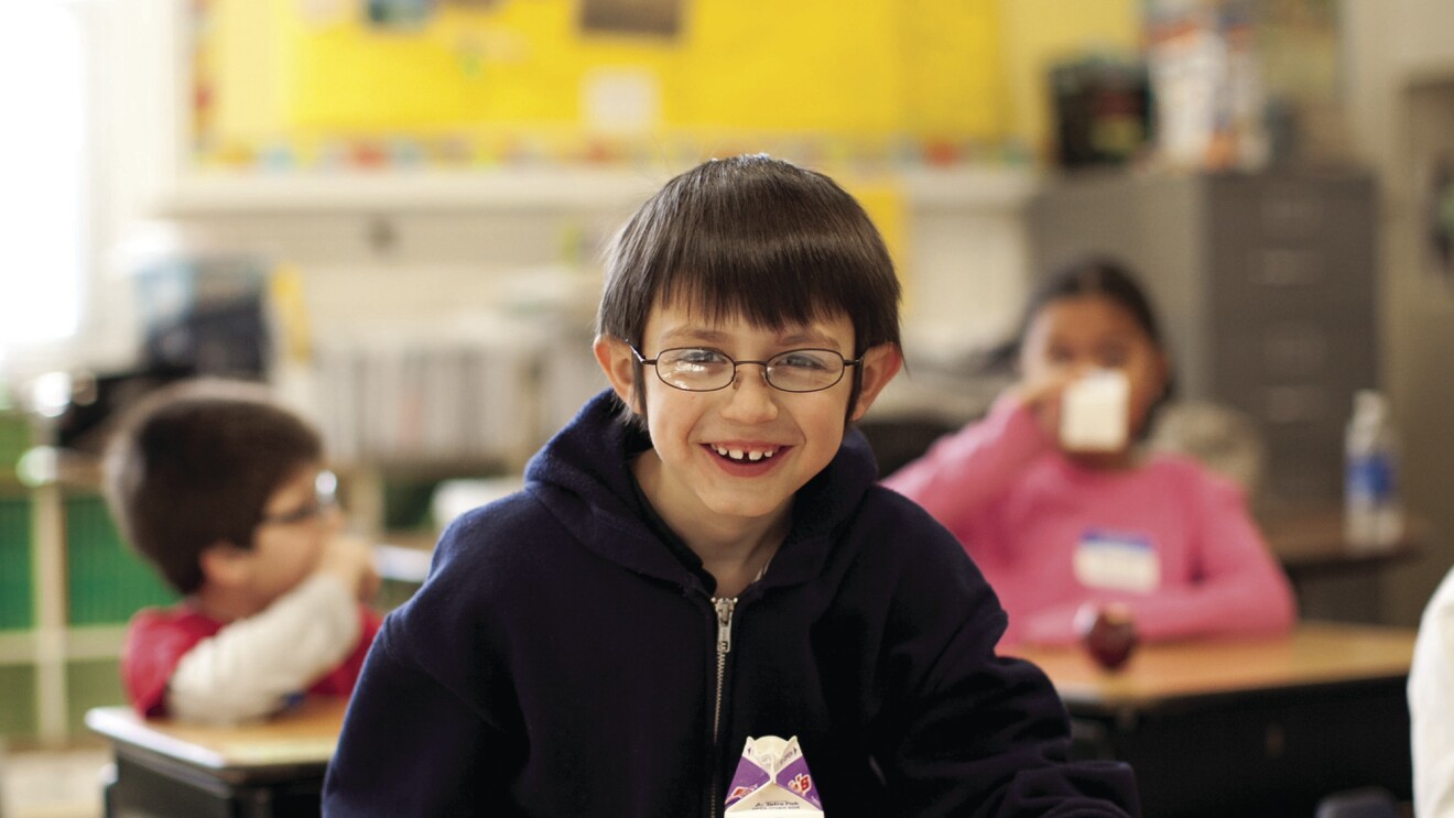 A boy sits at a school desk, smiling at the camera. On his desk are a carton of milk and a breakfast sandwich. Behind him, other students also eat and drink.