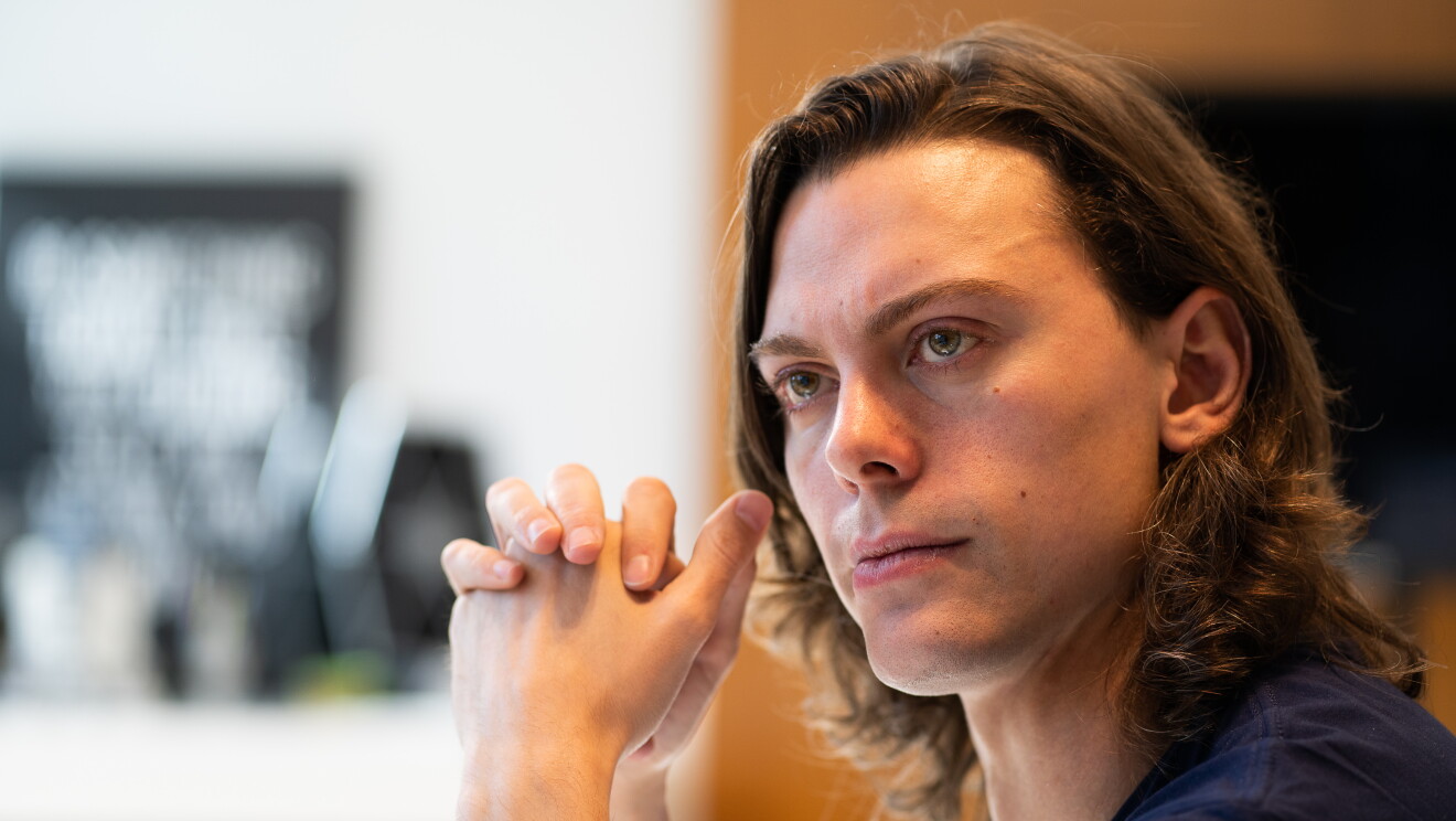 Joaquín Garralda, Responsable AWS de las cuentas de startups. Con el pelo largo y marrón, las manos entrecruzadas delante de la cara. Los ojos azules y una camiseta también azul. En primer plano está Joaquín y el fondo está desenfocado y es de color marrón, blanco y azul. 