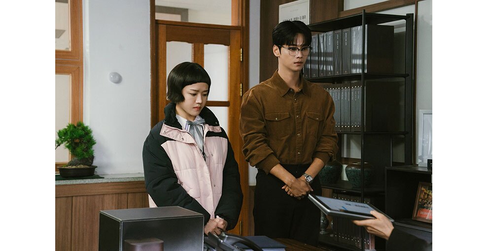 室内のドアの前に、2人の人物が並んで立っている。右側隅に、タブレットを持って2人に見せる手。室内にはファイルが入った棚や盆栽。