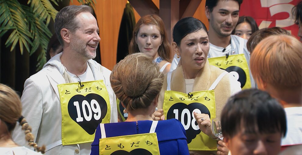 黄色いゼッケンを着けた人々が10人ほどたっている。ゼッケンには「ともだち」の文字と黒いハートマーク、その中に個別の番号。中央に青い服を着た人物の後ろ姿。左側に108番ゼッケンの人物が笑顔で、右側に109番ゼッケンの人物が戸惑いの表情で写っている。