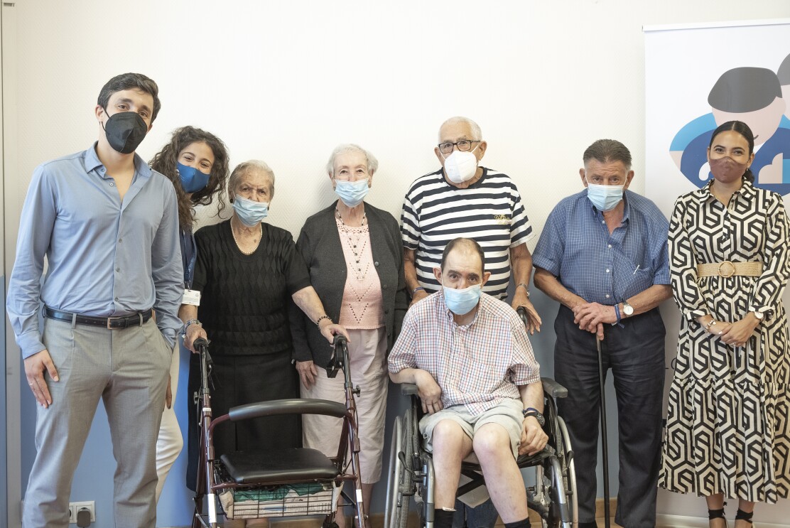 En la foto aparecen 8 personasm, cinco de ellas son mayores y tres son 1 chico y dos chicas jóvenes. De las personas mayores hay dos mujeres y tres hombres. Están todos de pie menos una persona mayor que va en silla de ruedas. Hay también una mujer mayor con caminador y un hombre con bastón.
Tiene una camiseta de manga corta, de rayas blanca y azul, gafas y el pelo blanco corto. Está mirando a cámara. Todos van con mascarilla. La primer personade la izquierda va con pantalón chino gris y camisa azul. Detrás de él aparece una chica con el pelo rizado marrón largo con un pantalón blanco. Al lado Hermelia con el pelo corto blanco y los ojos negros. Va con un collar de perlas, un jersey negro y unos pendientes también negros. Al lado otra señora con el pelo blanco, una chaqueta gris y un jersey de punto rosa. Una falda de color blanco hasta el tobillo. Antonio tiene una camiseta de manga corta, de rayas blanca y azul, gafas y el pelo blanco corto. Delante de él hay una persona en silla de ruedas con una camisa de manga larga y un pantalón contro. Y al lado de Antonio, con un bastón hay una persona mayor con una camisa azul de manga corta y un pantalón también azul. Y al final de la foto aparece la última chica joven con el pelo negro recogido en una cola baja. El vestido es de color beige con figuras geométricas negras y un cinturón.
