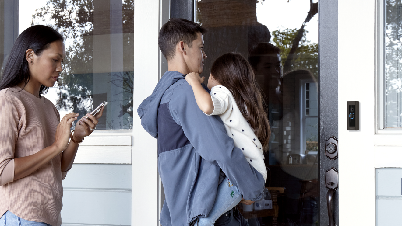 An image of a family walking into their house. A man is holding a child while opening the door and a woman is close behind him using her phone.