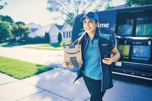 A smiling Amazon delivery driver walks carrying a package to deliver. The delivery vehicle is parked on the street.