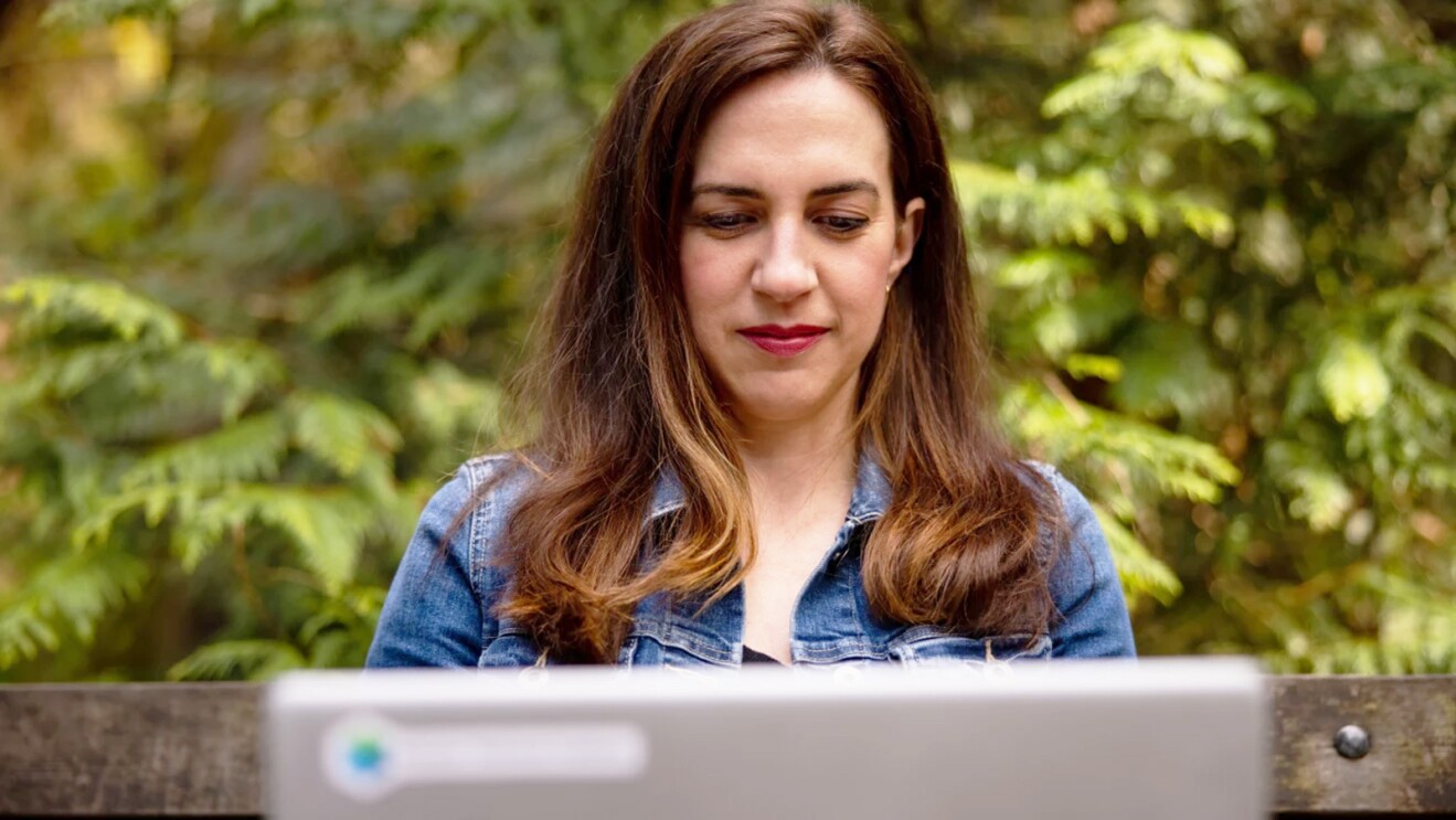 Ashely Rajagopal sits on a bench outside with vibrant greenery behind her. She is focused while she works on her laptop.