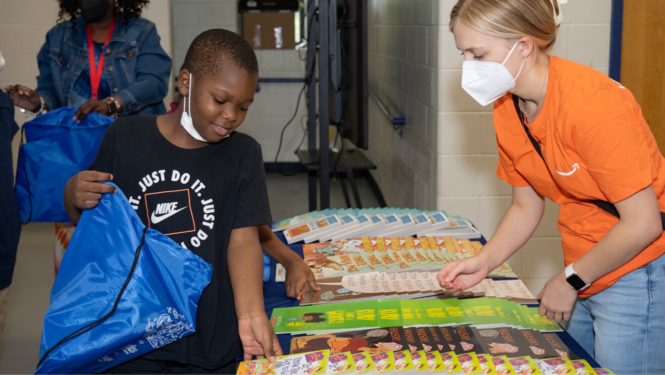 An image of an Amazon employee volunteer wearing an orange shirt and a student holding a bag while picking books placed on a table.  