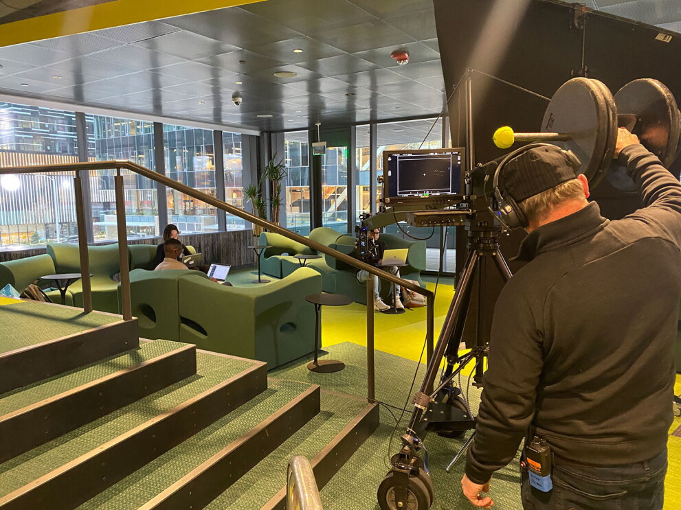 An image of a film crew filming a group of people sitting in the lobby of an Amazon office building on their laptops.