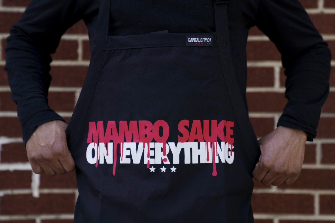 A person wearing a black apron that reads, "Mambo Sauce on everything."