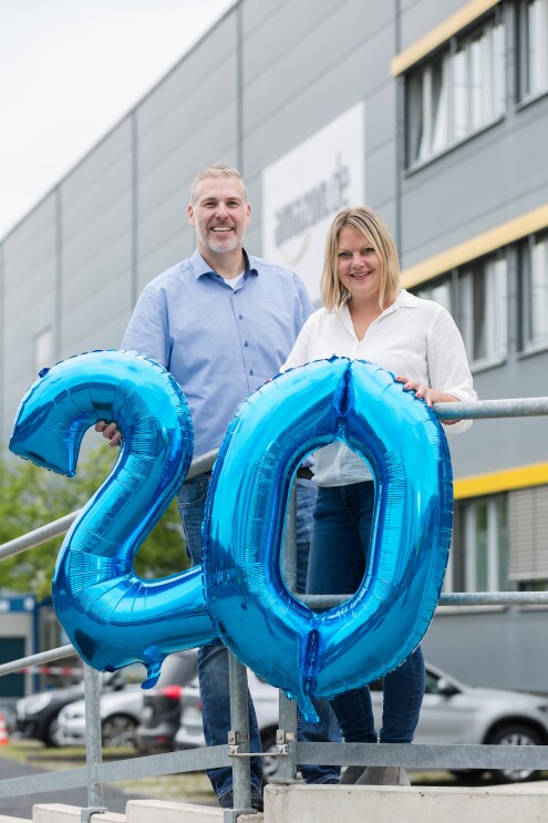 Ein Mann in blauem Hemd und Jeans und eine blonde Frau in weißer Bluse und Jeans stehen lächelnd vor einem Logistikzentrum. An der Fassade ist das Amazon.de Logo erkennbar. Beide halten blaue, aufgeblasene Zifffern - 2 und 0 - vor sich.