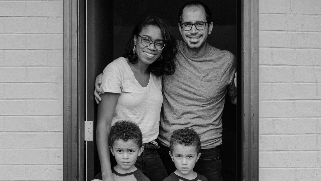 A black and white image of a family smiling for a photo while standing n the doorway of a brick home. There is a mother and father and two sons.