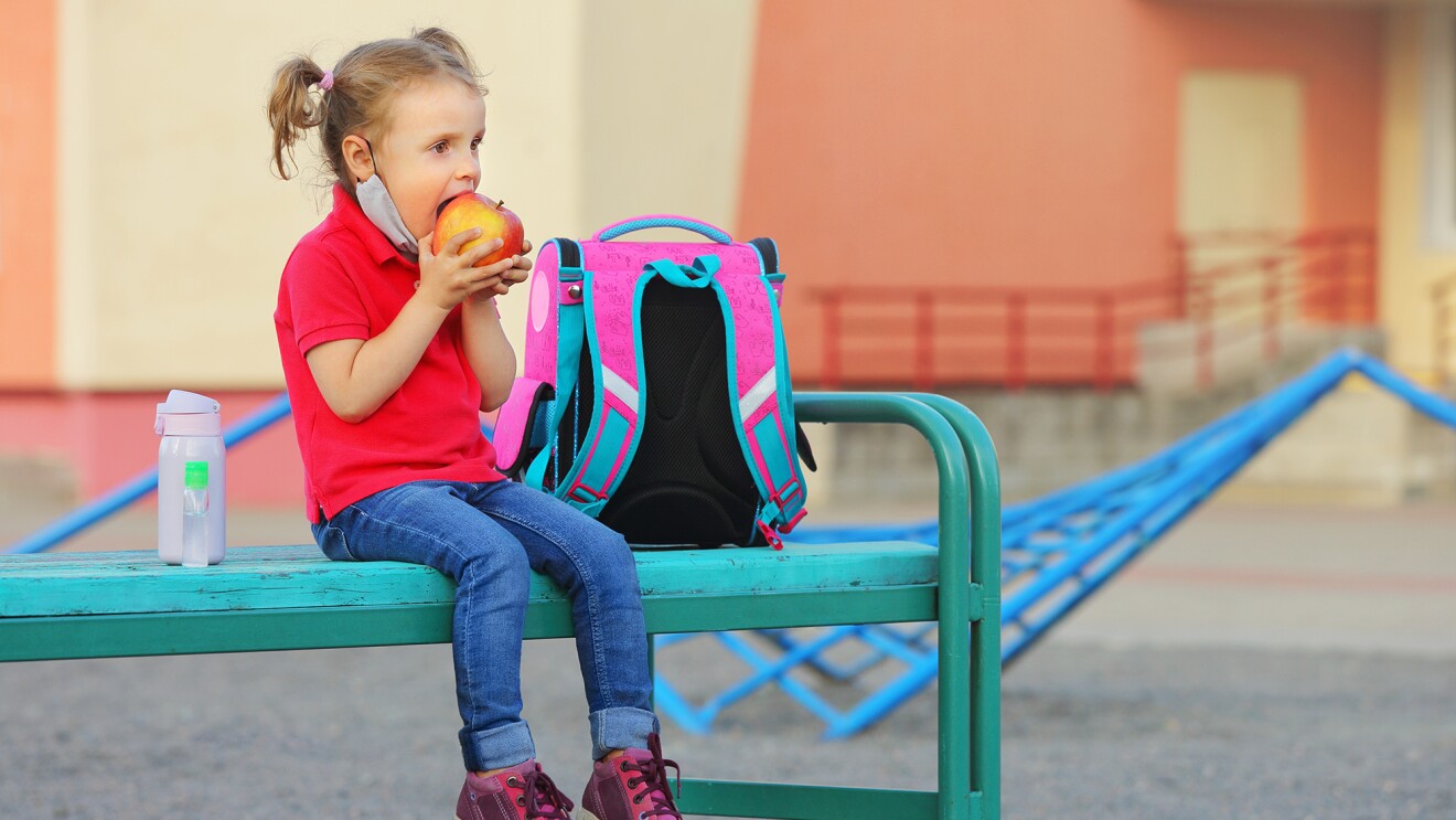 A school-age child sits on a bench in a school yard. She has pulled her mask down and is taking a bite of an apple that she holds in her hands .