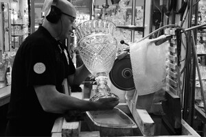 Un artisan est en train de polir un vase de cristal sur une machine électrique. Il porte un casque de protection sur les oreilles. Au fond on aperçoit des vitrines de créations en cristal. C'est une photo noir et blanc.