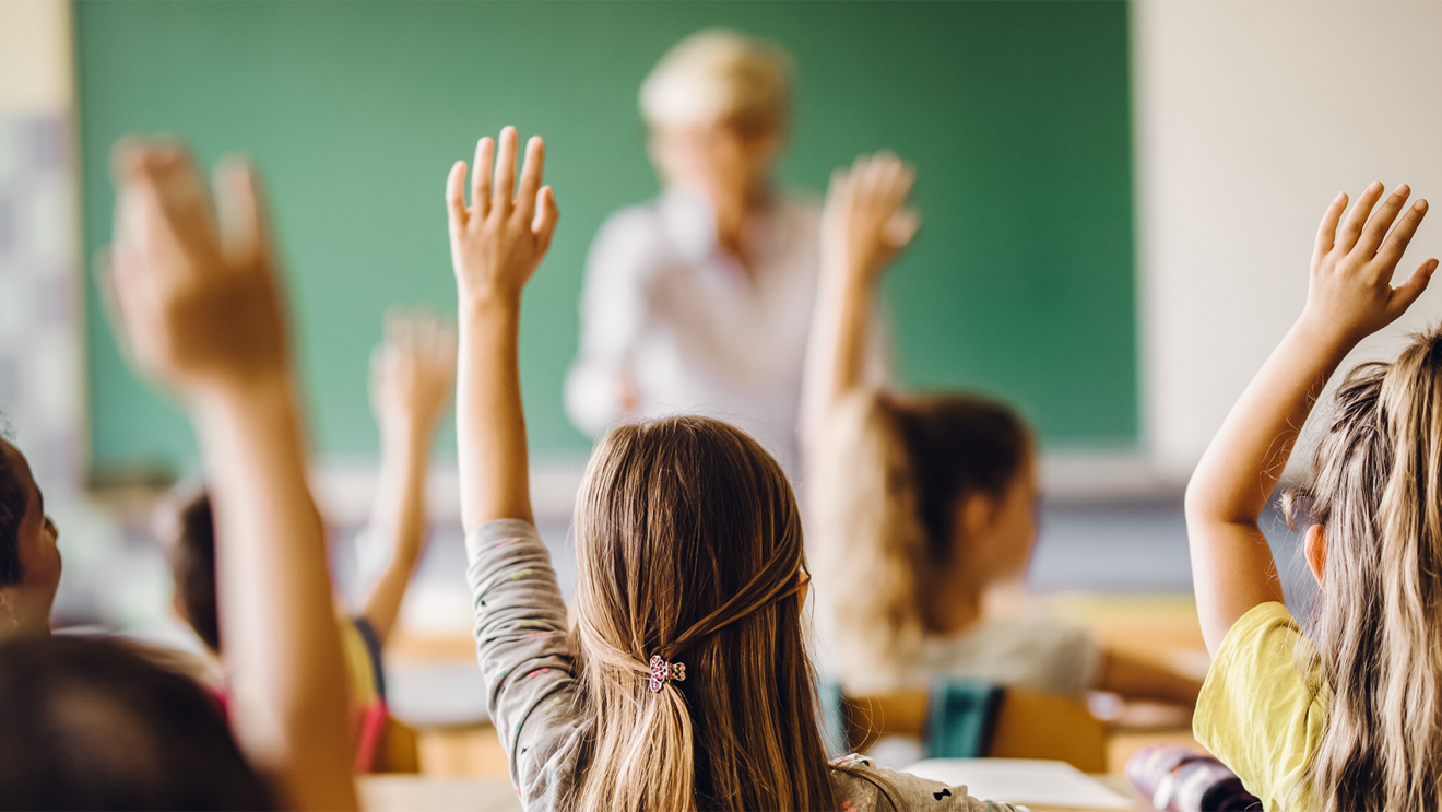 Elementary age students sit in a classroom. Many raise one hand to signify they'd like the teacher to choose them. 