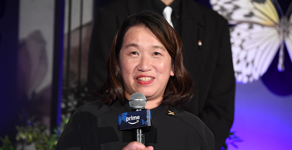 大きな蝶のモチーフや青い花が設置された青い壁を背に、マイクを持って話す女性。黒い服を着用。