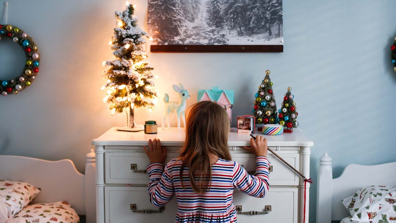 A young girl stands in front of a dresser, in front of an Alexa Echo device and small Christmas tree. 