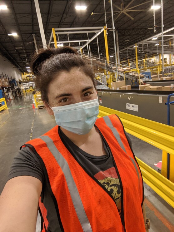 Sarah Freeman, an Amazon associate, is smiling for a selfie inside the Amazon fulfillment center where she works. She is wearing a mask and other Amazon safety gear like an orange vest. 