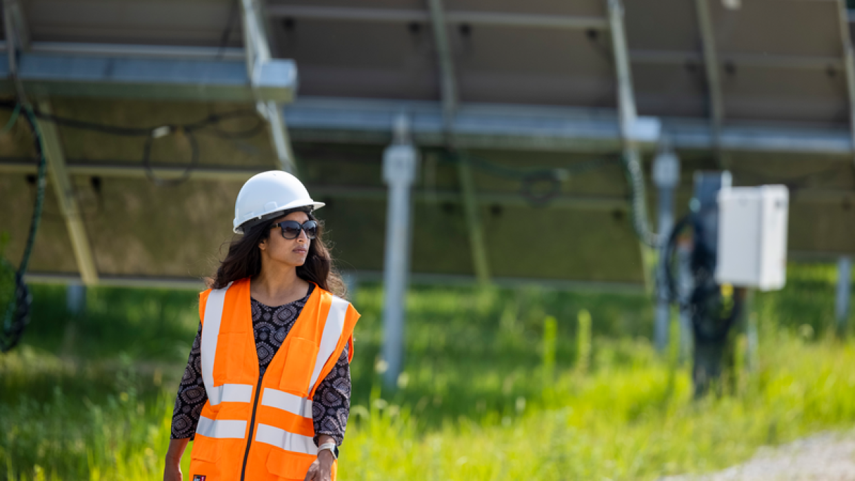 En una granja solar, delante de la parte trasera de tres placas aparece caminando una mujer con gafas de sol, casco blanco, y el pelo ondulado largo. Lleva una jeans, un chaleco de color naranja con un jersey negro estampados con formas geométricas grises.