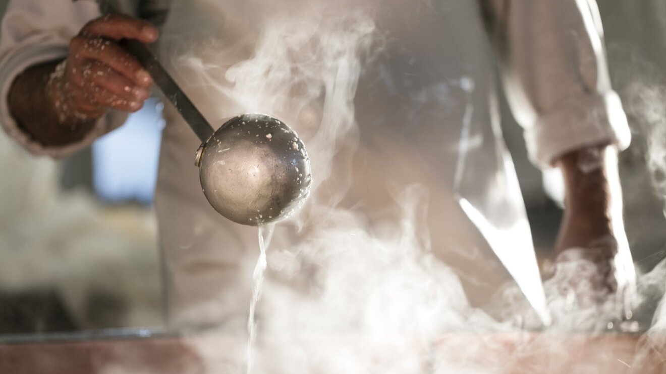 Primo piano di un mestolo retto da un uomo che lavora alla cottura della pasta del formaggio nella Latteria Due Madonne. L'uomo regge il mestolo con la mano sinistra e solleva lo strumento in mezzo al fumo che sale dalla pentola.
