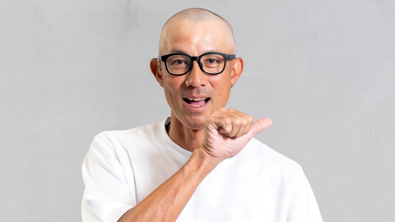 白いTシャツを着た男性が顔の前に親指だけを広げほかの指は握った形を作っている。口元は開いている