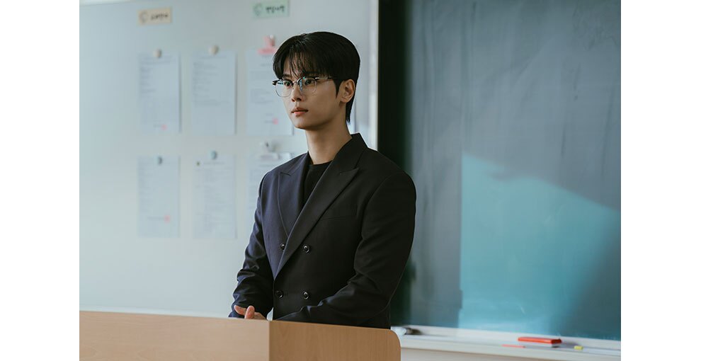 黒板の前、教壇に向かって立つ眼鏡の男性。教壇の上で手を組んでいる。