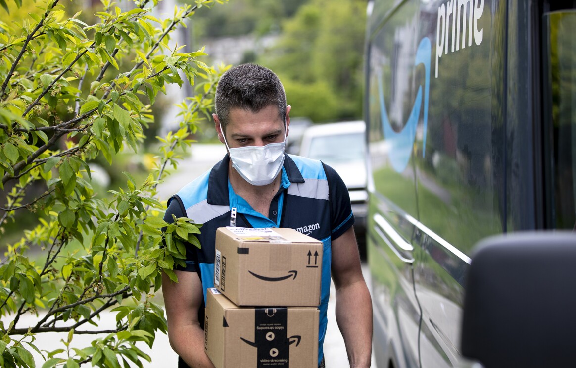 A delivery driver wearing a face mask walks alongside an Amazon Prime delivery van, carrying packages.