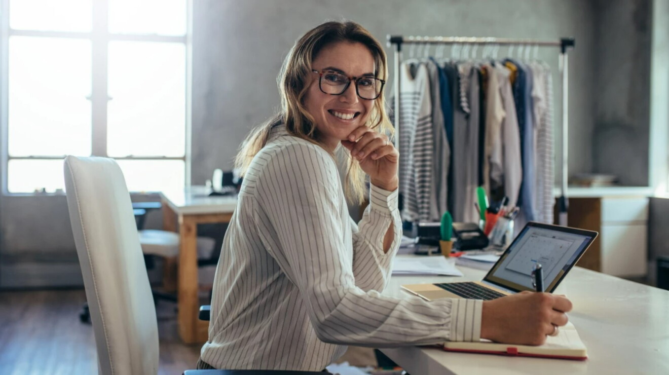 Female business owner sits at her home studio desk with notepad and laptop