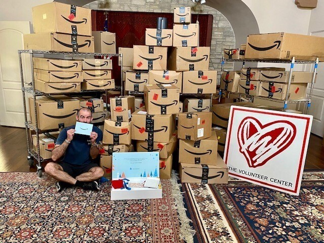 An image of Amazon boxes stacked up with a man sitting in front of the boxes holding the Delivering Smiles invitation for charities selected for this year's campaign. There's also a sign with a big heart and "The Volunteer Center" on it.