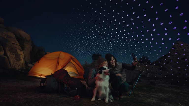 Two people and a husky sitting by an illuminated orange tent during nighttime camping
